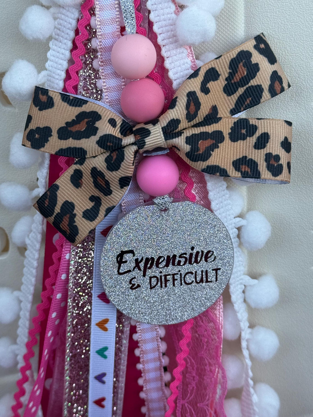 Close-up of leopard print bow and glitter Expensive & Difficult charm disc with layered hot pink, silver glitter, lace, and heart ribbon tassel on a silicone beaded bag charm
