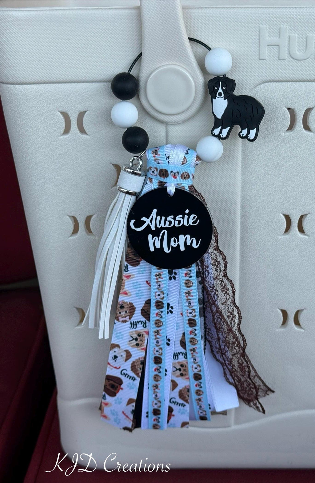 Close-up of Aussie Mom silicone beaded bag tassel clipped to white Hurley tote bag handle showing alternating black and white round silicone bead loop with black and white Australian Shepherd silicone dog charm, black round Aussie Mom acrylic pendant with white script, and cascading light blue dog-print grosgrain, white leather fringe, brown grosgrain, and black lace ribbon tassel, KJD Creations dog mom bag charm