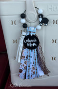 Close-up of Aussie Mom silicone beaded bag tassel clipped to white Hurley tote bag handle showing alternating black and white round silicone bead loop with black and white Australian Shepherd silicone dog charm, black round Aussie Mom acrylic pendant with white script, and cascading light blue dog-print grosgrain, white leather fringe, brown grosgrain, and black lace ribbon tassel, KJD Creations dog mom bag charm