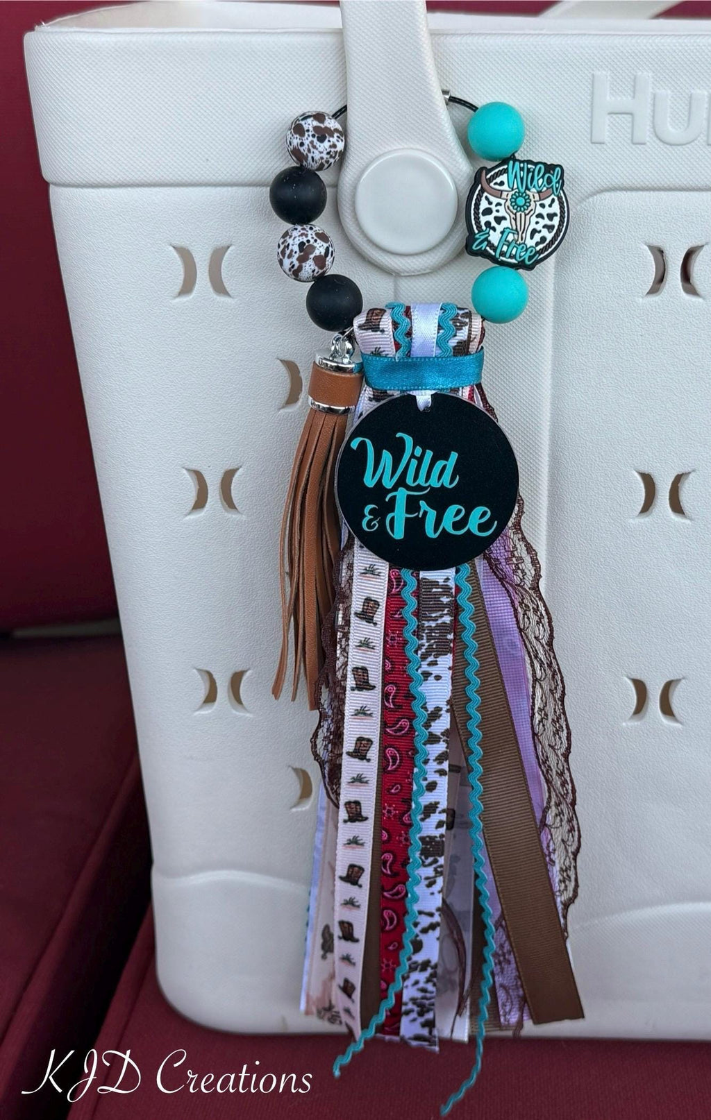 Western cowgirl bag tassel with Wild & Free charm disc, mixed ribbon fringe, leather tassel, and silicone beads clipped to a white Bogg-style bag