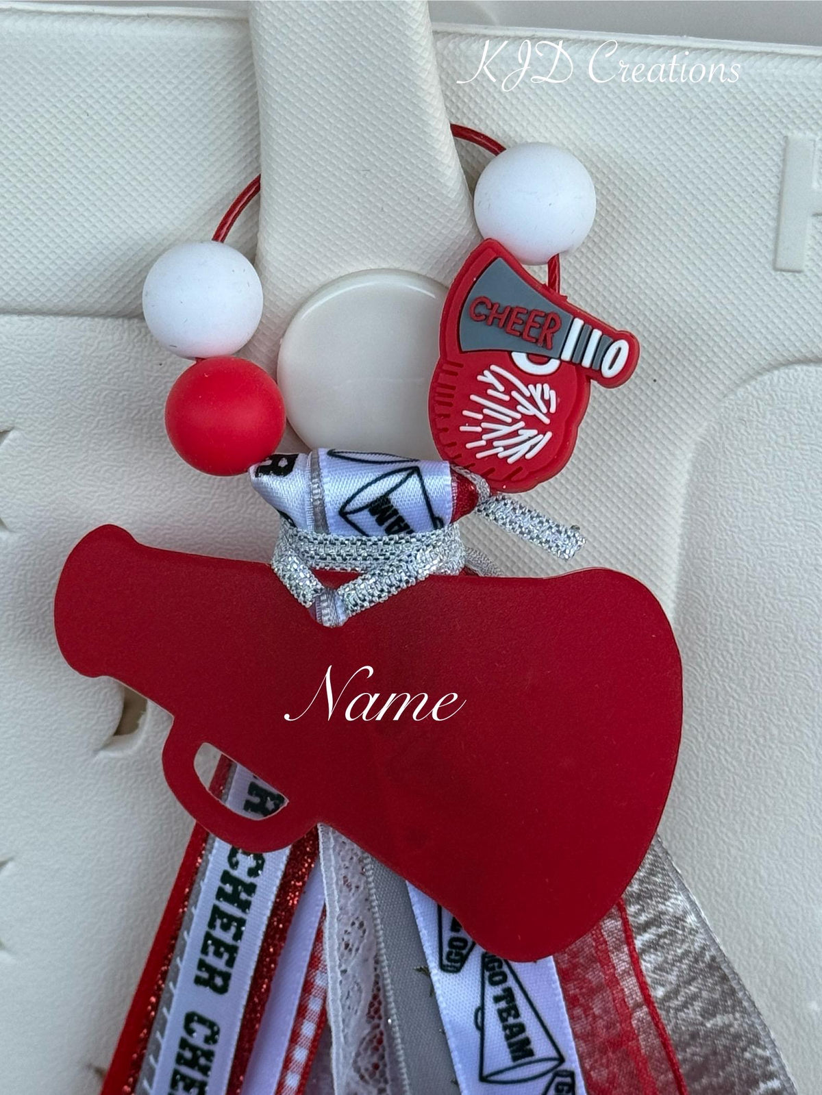 Close-up of red personalized cheer silicone beaded bag tassel showing wire loop with red and white silicone beads and red Cheer 110 megaphone silicone charm, silver glitter ribbon bow, and large red acrylic megaphone-heart pendant with personalized name in white script, KJD Creations cheerleading bag charm detail