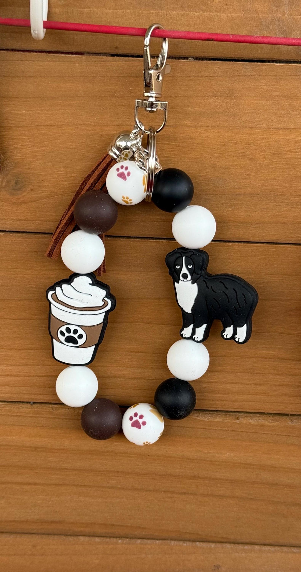 A beaded keychain or bag charm with white, black, and brown beads, featuring a small dog figure, a coffee cup with a paw print, and paw-print details, hanging against a wooden background.