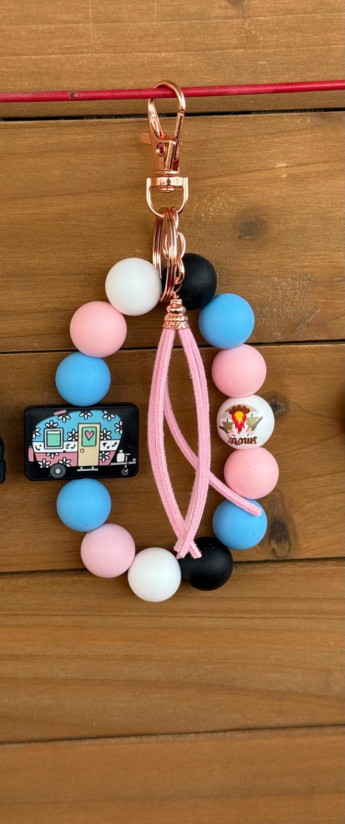 A beaded keychain or phone strap charm with pastel pink, blue, white, and black silicone beads, a pink braided cord, a rose gold clasp, and a small decorative charm, shown on a wooden surface.