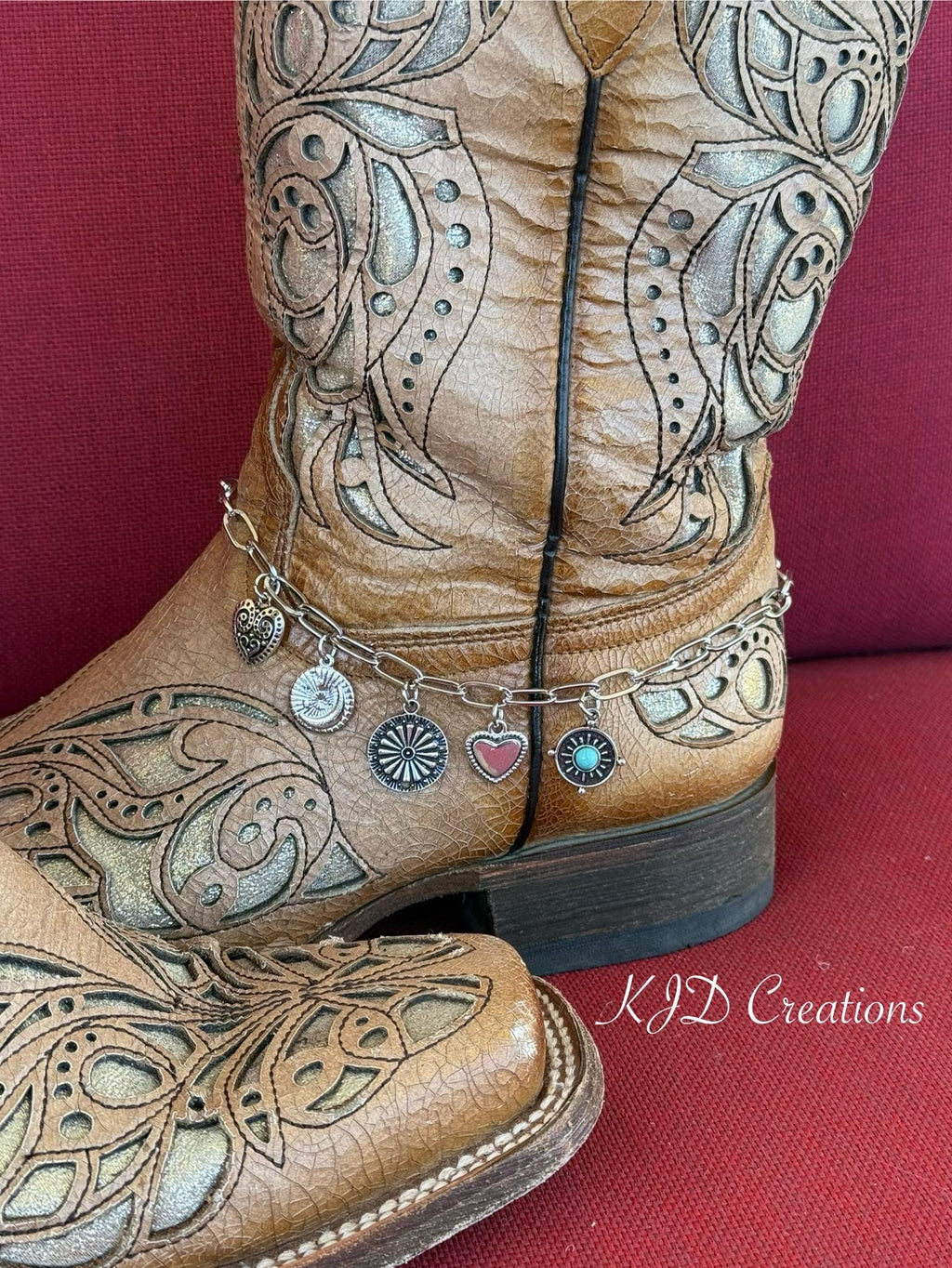 A close-up of a brown tooled leather cowboy boot with ornate western embroidery ankle with silver paperclip chain boot bracelet featuring five dangling charms: silver filigree heart, sunburst coin, daisy wheel concho, silver heart, and turquoise stone concho disc