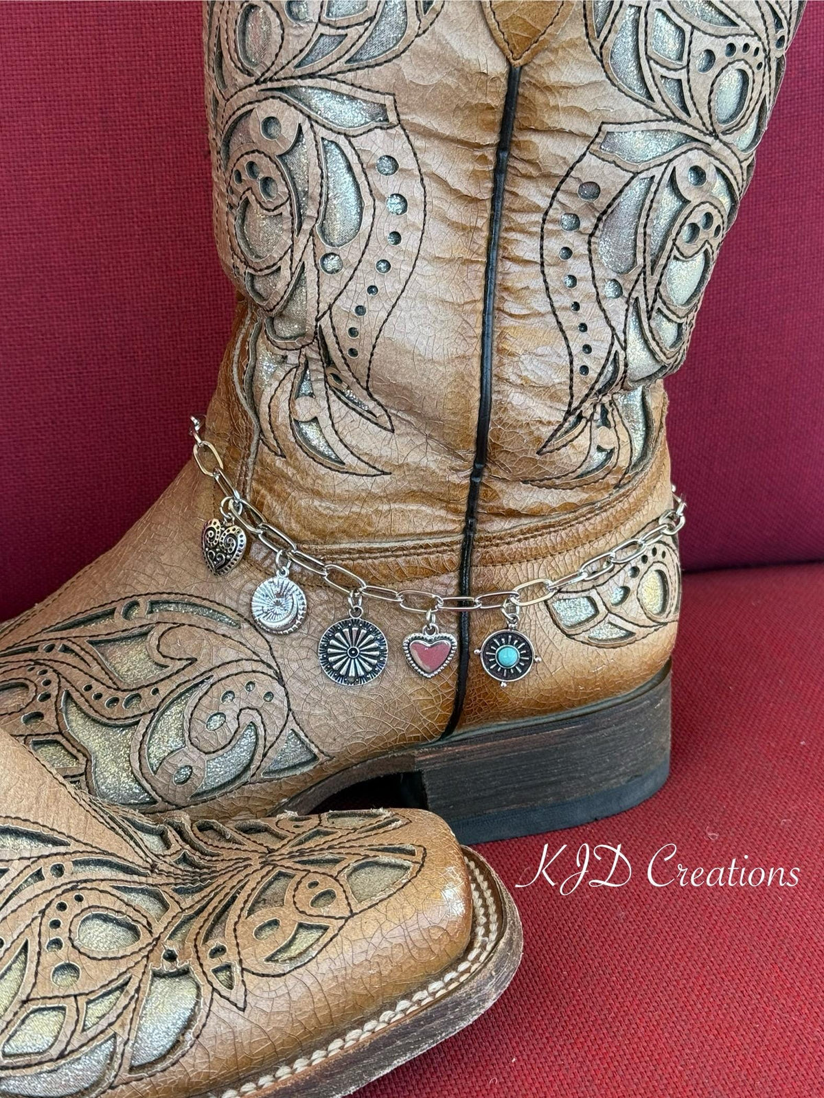 A close-up of a brown tooled leather cowboy boot with ornate western embroidery ankle with silver paperclip chain boot bracelet featuring five dangling charms: silver filigree heart, sunburst coin, daisy wheel concho, silver heart, and turquoise stone concho disc