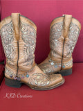 A pair of tan western cowboy boots against red background wearing silver paperclip chain boot bracelets with five dangling charms including silver filigree heart, sunburst coin, daisy wheel concho, silver heart, and turquoise stone concho disc