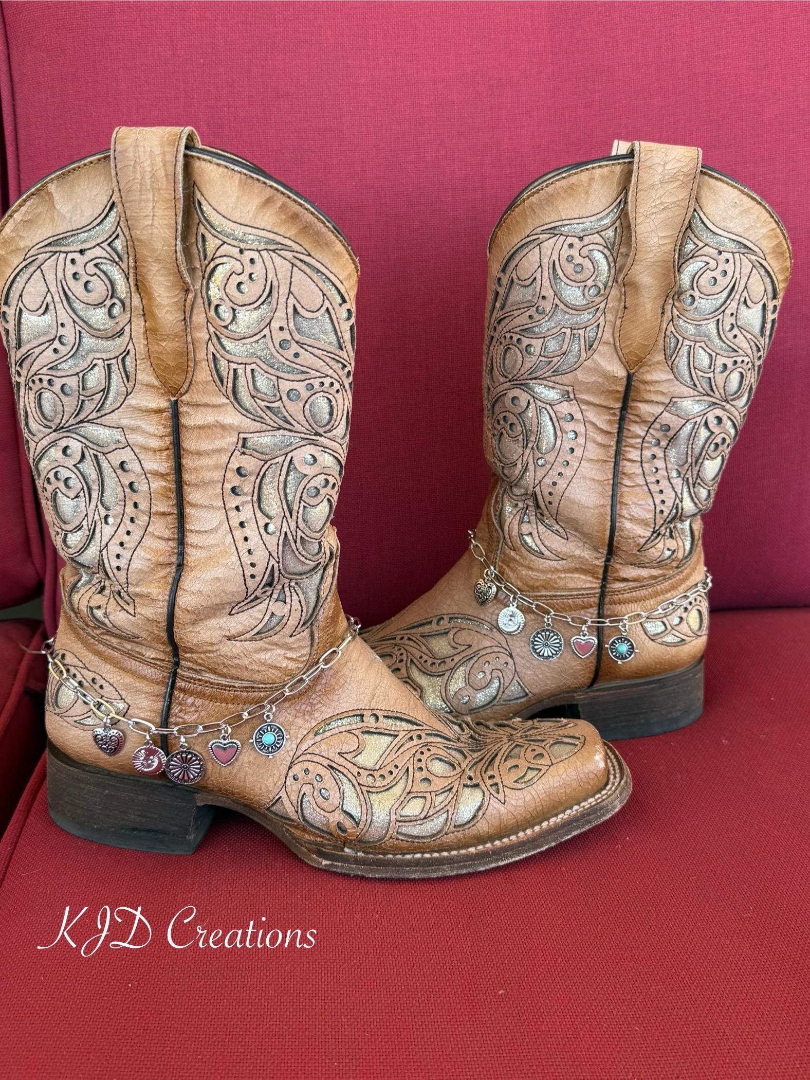 A pair of tan western cowboy boots against red background wearing silver paperclip chain boot bracelets with five dangling charms including silver filigree heart, sunburst coin, daisy wheel concho, silver heart, and turquoise stone concho disc