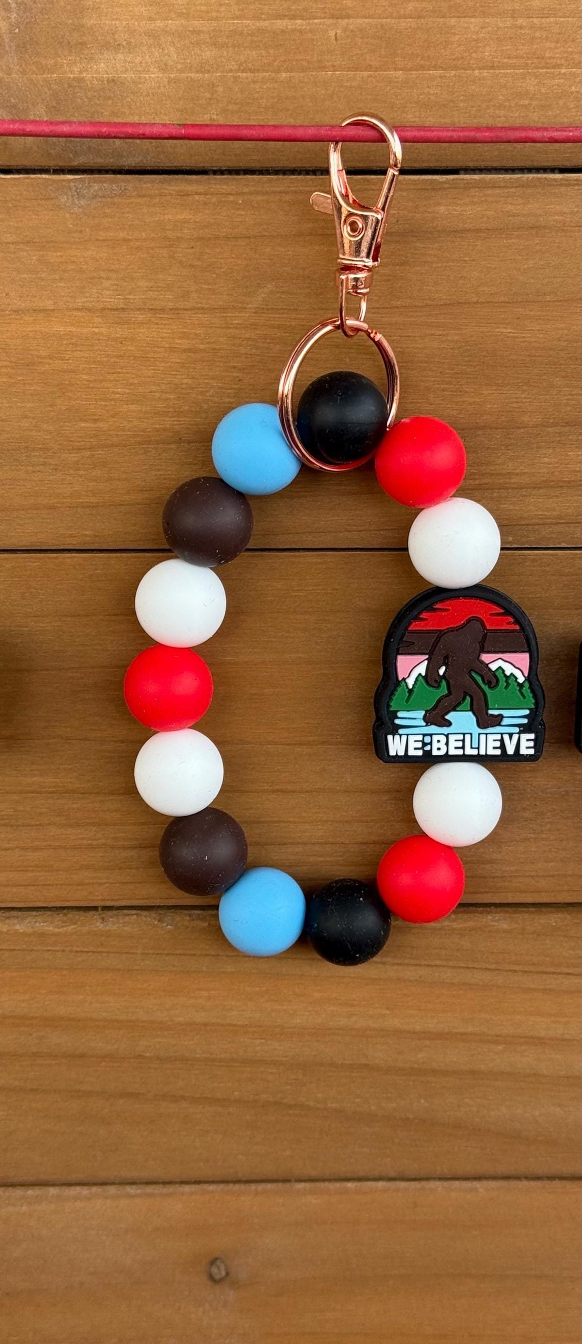 A beaded keychain with alternating red, white, blue, brown, and black beads, featuring a rose gold clasp and a small 'We Believe' Bigfoot patch, displayed on a wooden background.