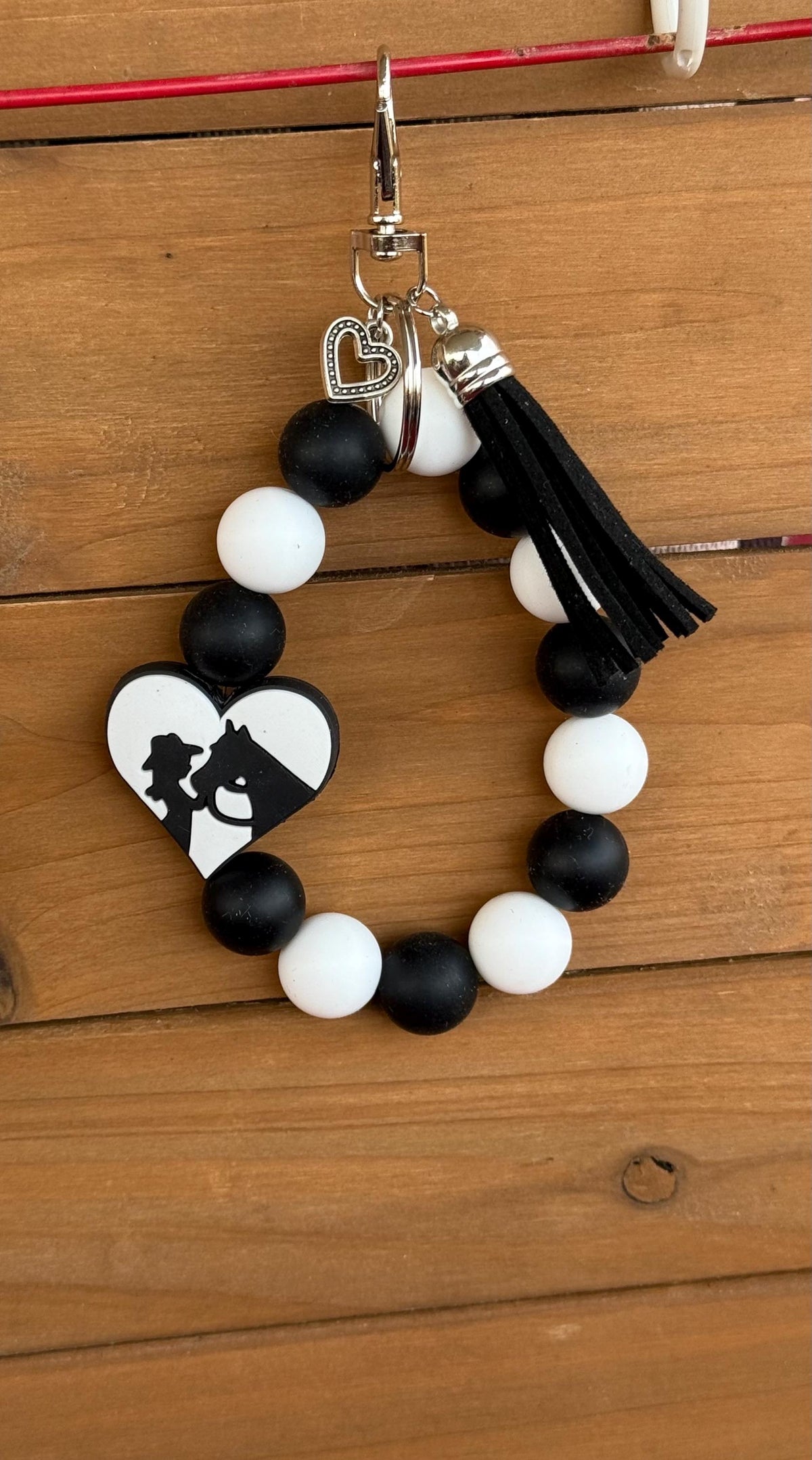 A beaded keychain or bag charm with alternating black and white round beads, a black tassel, a silver clasp, and a heart-shaped charm featuring a black silhouette of cowgirl and Horse on a white background. The keychain is displayed hanging from a red string against a wooden plank background.