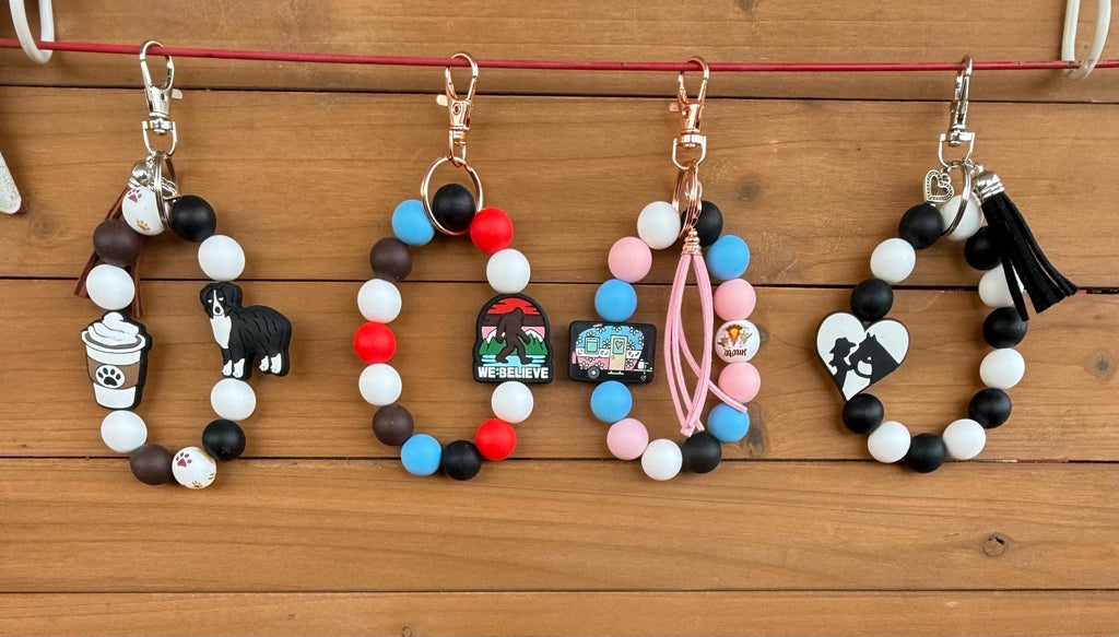 Four beaded keychains with charms are displayed on a wooden surface. The keychains feature black, white, and colored beads, metal clips, and various charms including a dog figure, a coffee cup, a small landscape piece, pink tassels, and a heart with a black-and-white portrait.