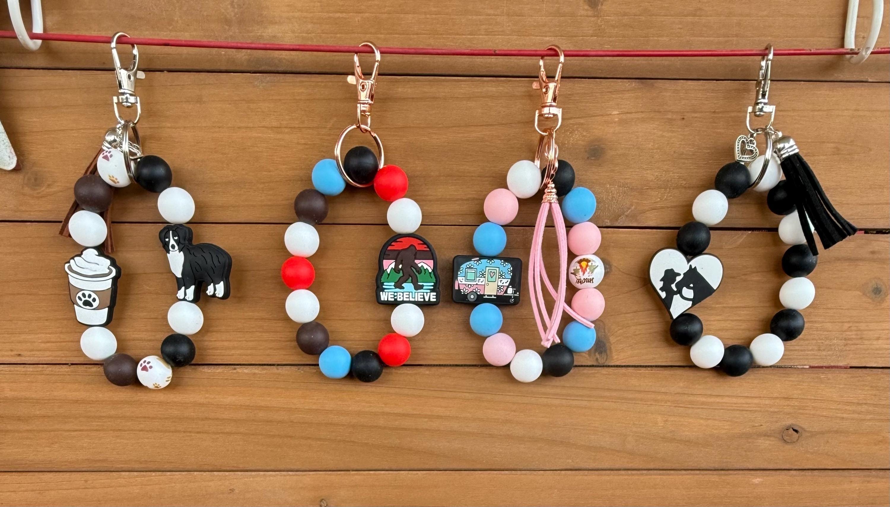Four beaded keychains with charms are displayed on a wooden surface. The keychains feature black, white, and colored beads, metal clips, and various charms including a dog figure, a coffee cup, a small landscape piece, pink tassels, and a heart with a black-and-white portrait.