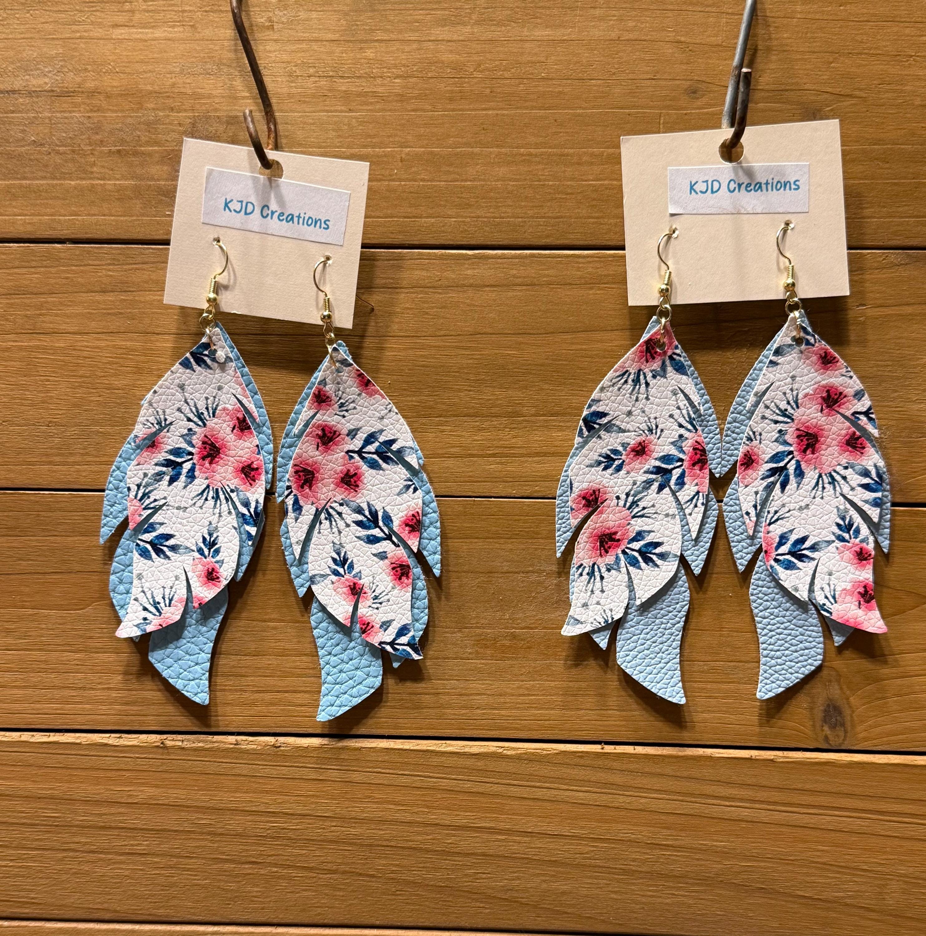 A pair of dangling leaf-shaped earrings with a floral print, hanging from kraft paper tags labeled KJD Creations on a wooden surface.
