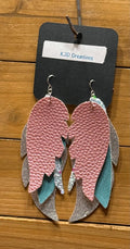 A pair of handmade feather-shaped dangle earrings displayed on a black card labeled 'KJD Creations' against a wooden background. The earrings feature layered pink textured faux leather, teal blue fabric, and silver glitter feathers on silver hooks.