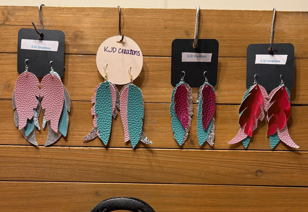 Four pairs of handmade feather-style dangle earrings displayed on black tags against a wooden wall. The earrings feature layered feather shapes in pink, teal, magenta, and silver, with two tags reading KJD creations.