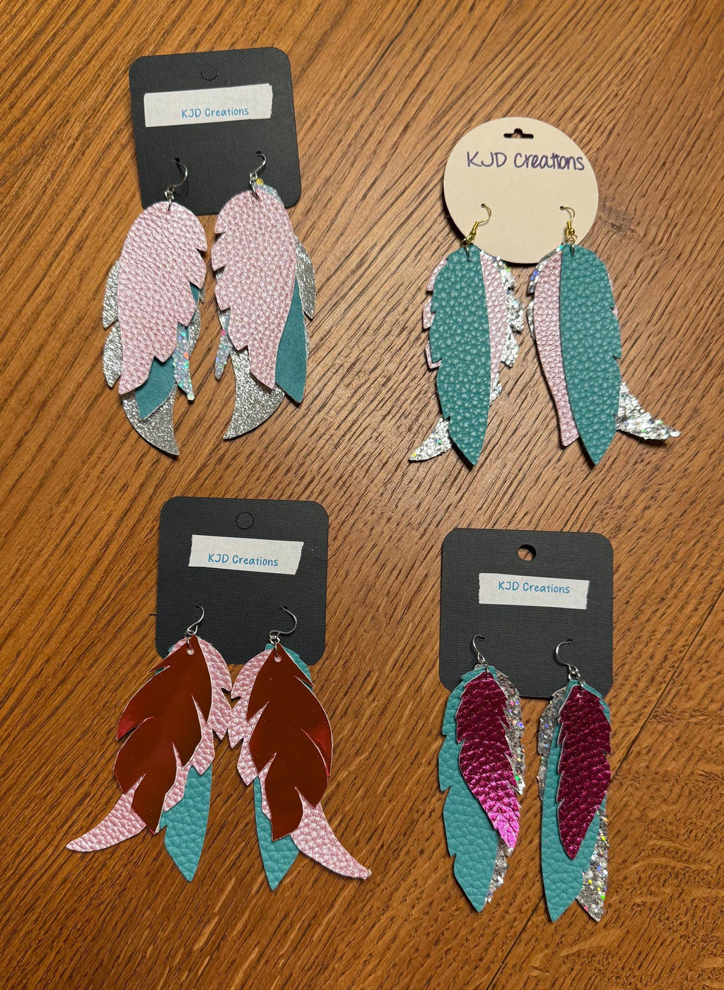 Four pairs of 3D feather drop earrings displayed on black and kraft paper cards on a wooden table. The earrings feature textured leather or faux leather feathers in pink, teal, brown, and magenta with silver glitter accents.