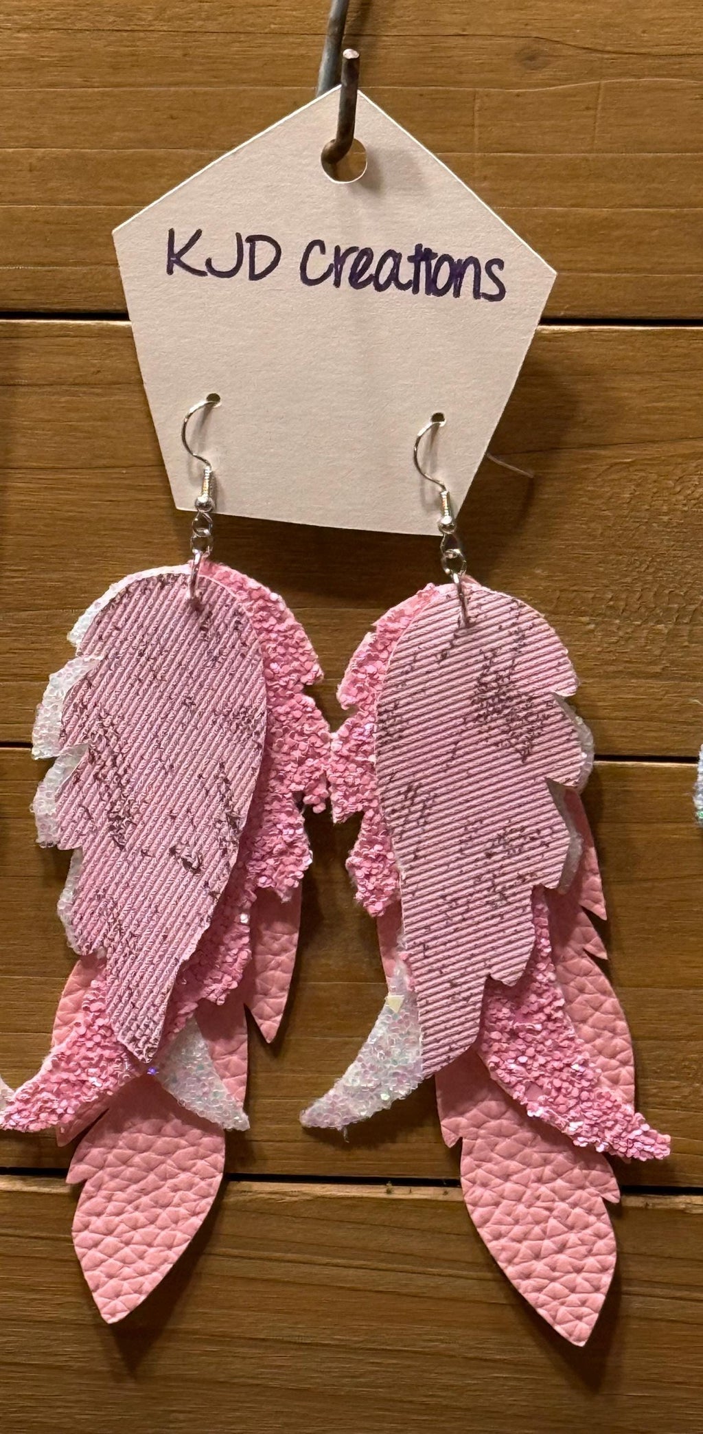 Pink textured leather angel wing drop earrings hanging from silver hooks on a white paper tag that reads 'KJD creations' against a wooden background.