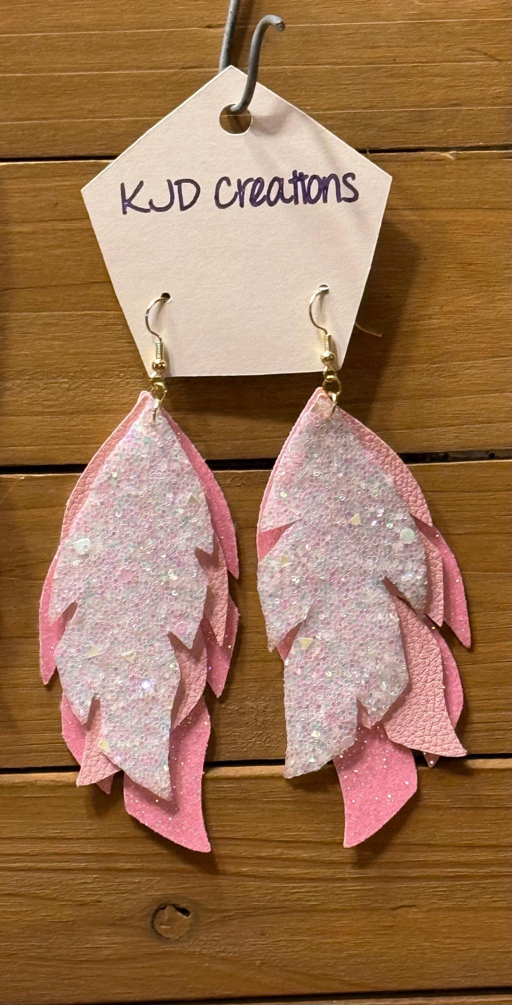 Pink feather-shaped dangle earrings with glittery silver-white centers, hanging from a beige display card labeled 'KJD creations' against a wooden background.