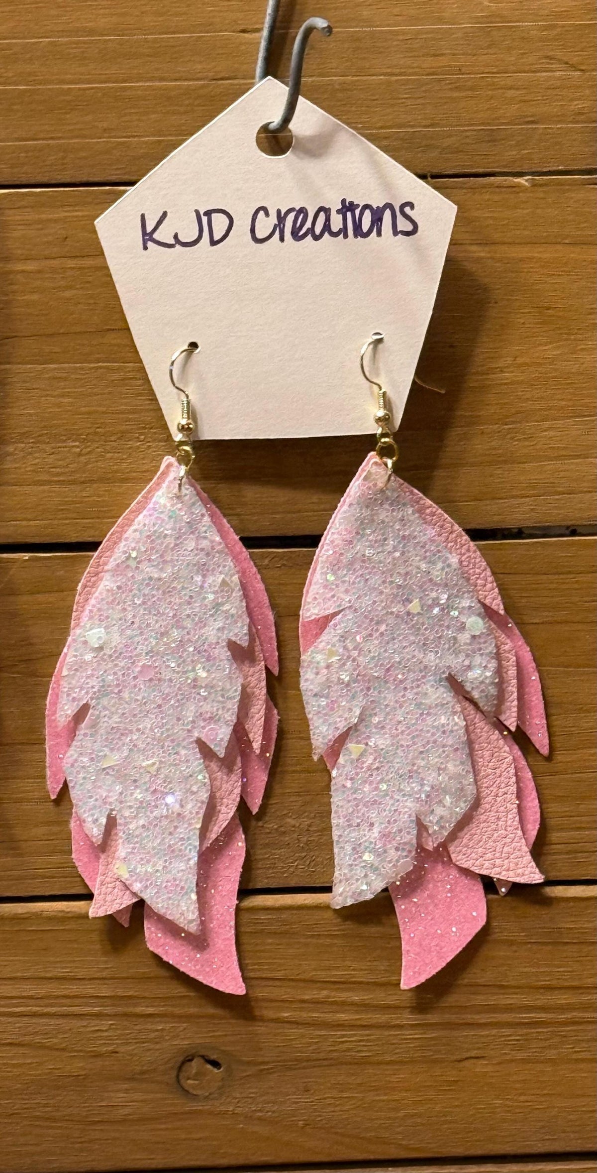 Pink feather-shaped dangle earrings with glittery silver-white centers, hanging from a beige display card labeled 'KJD creations' against a wooden background.