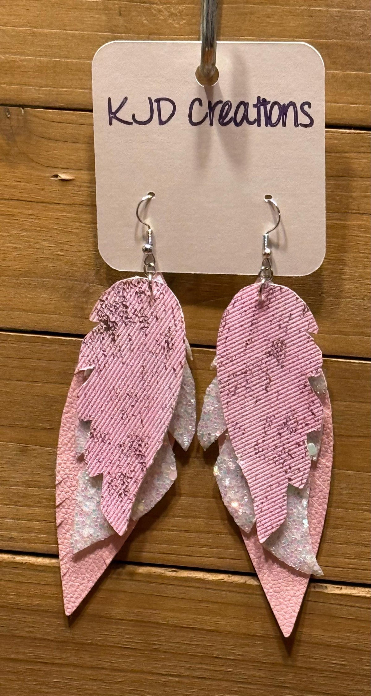 Pink feather-shaped dangle earrings with layered textured and glittery pink and white pieces, hanging from silver hooks on a display card that reads KJD Creations, set against a wooden background.
