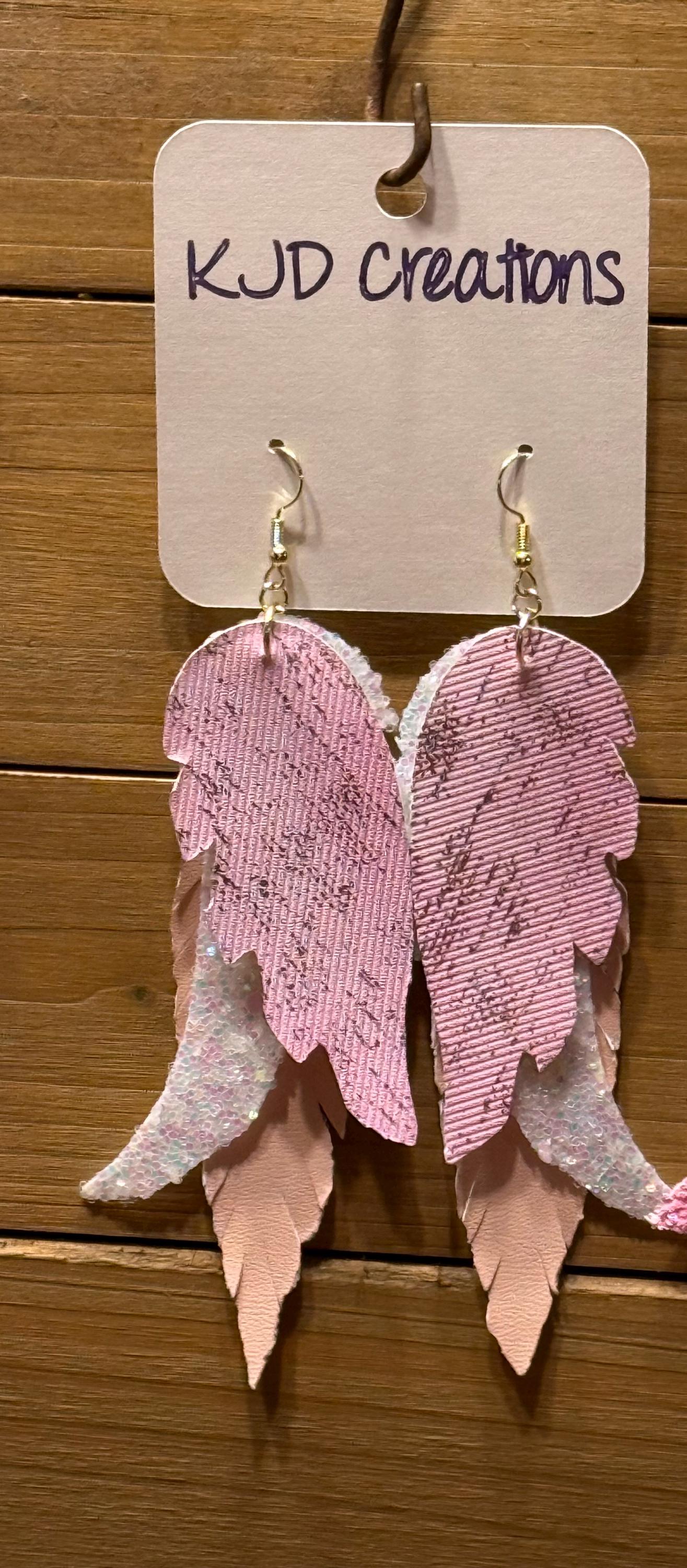 Pink and white feather wing dangle earrings with gold fishhook wires, hanging from a white display card that reads 'KJD creations' against a wooden background.
