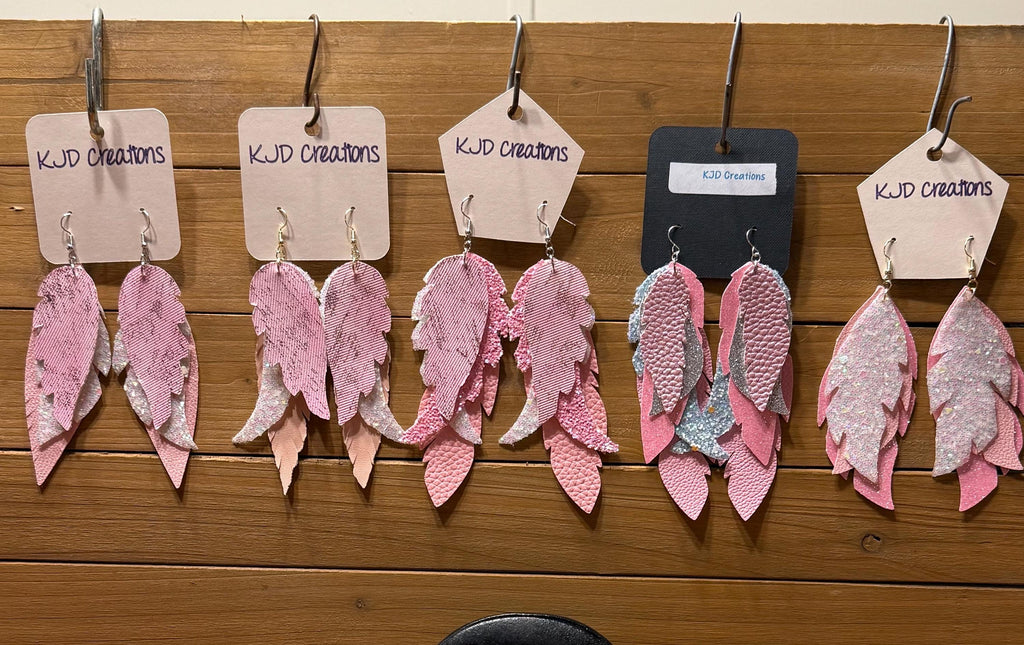Five pairs of handmade feather-shaped dangle earrings in various pink and silver patterns, hanging from metal hooks on a wooden wall. Each pair has a paper tag with 'KJD creations' written on it, except for one pair with a black tag labeled 'ECO Charlies'.