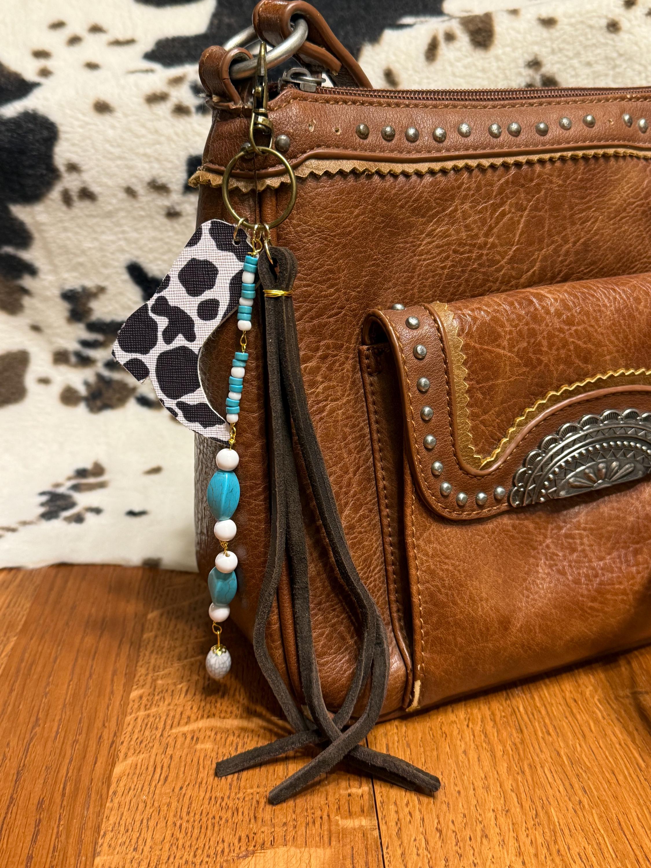 Front view of brown studded Western leather crossbody bag on wood floor against cowhide rug background with boho Western bag charm featuring black and white cow print faux leather cowboy boot charm, turquoise heishi and oval bead strand with white round beads, and dark brown leather fringe tassel