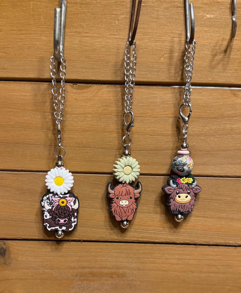 Three highland cow tumbler dangle charms hanging on a display rack — White Daisy with cow print charm, Green Daisy with pink highland cow charm, and Cow Floral Bead with floral bead and highland cow charm, all on silver chains by KJD Creations