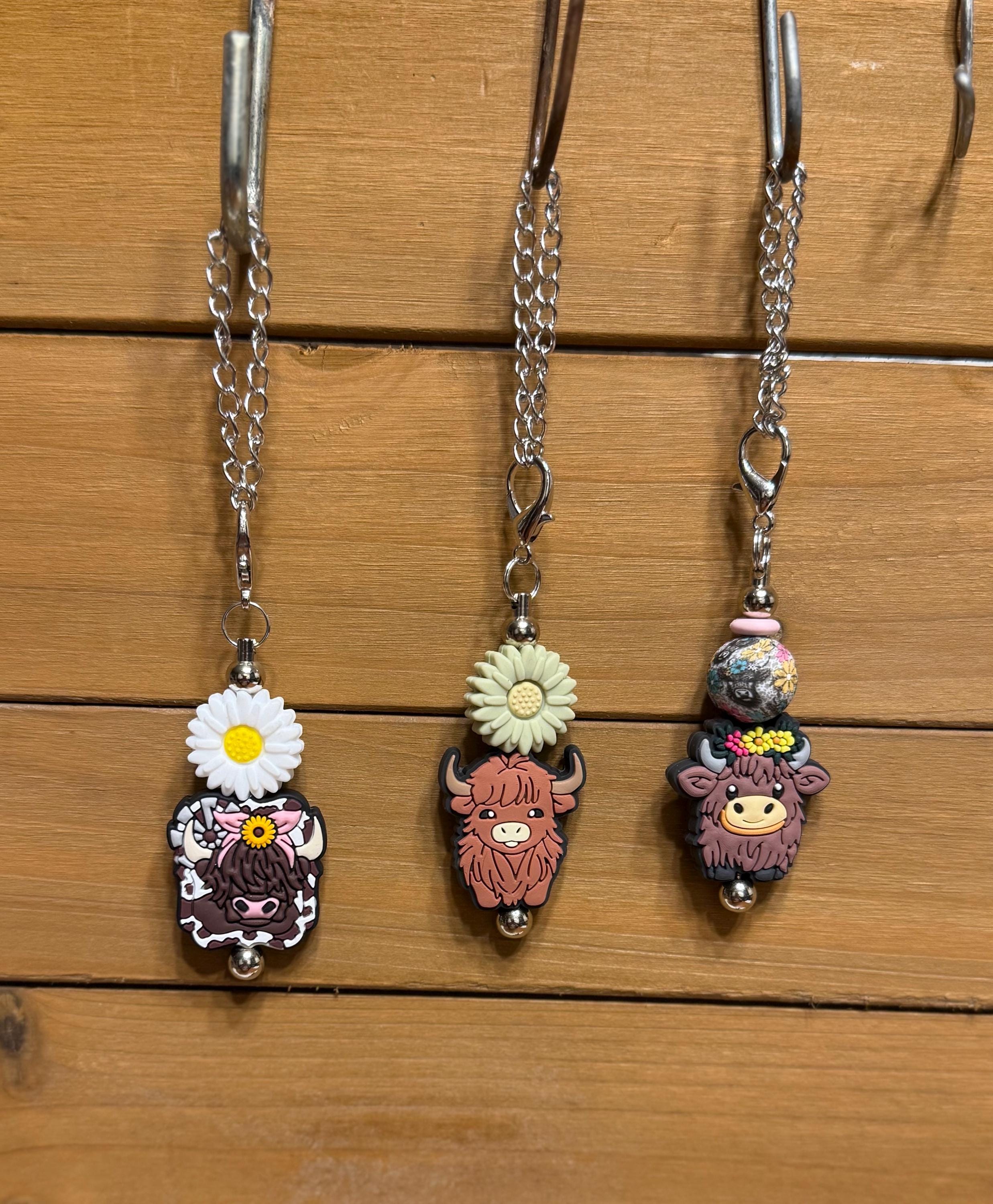 Three highland cow tumbler dangle charms hanging on a display rack — White Daisy with cow print charm, Green Daisy with pink highland cow charm, and Cow Floral Bead with floral bead and highland cow charm, all on silver chains by KJD Creations