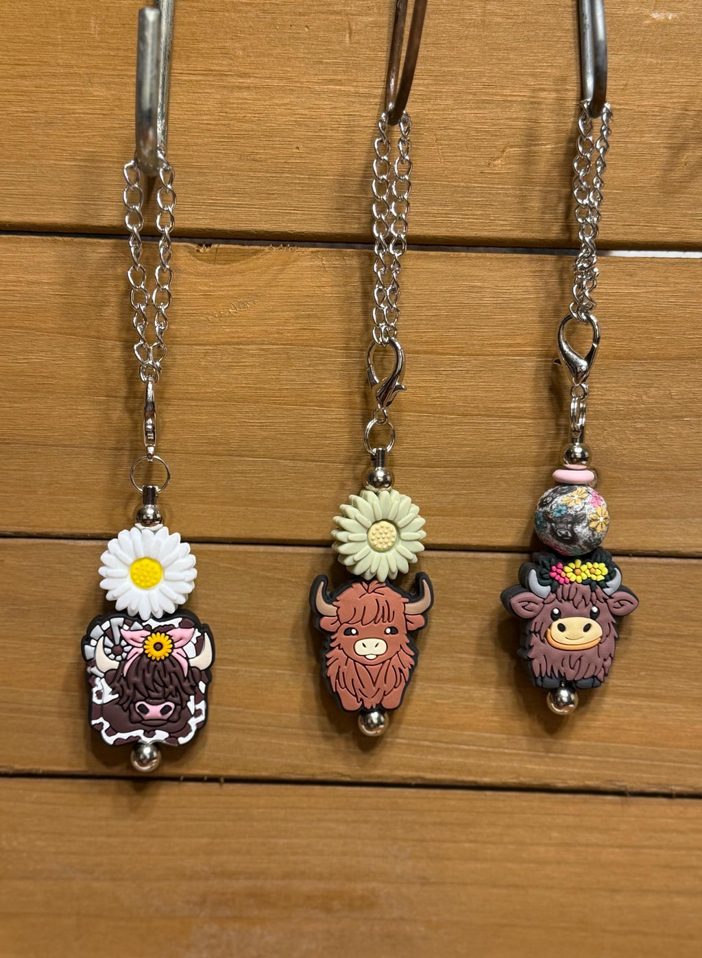 Three highland cow tumbler dangle charms hanging on a display rack — White Daisy with cow print charm, Green Daisy with pink highland cow charm, and Cow Floral Bead with floral bead and highland cow charm, all on silver chains by KJD Creations