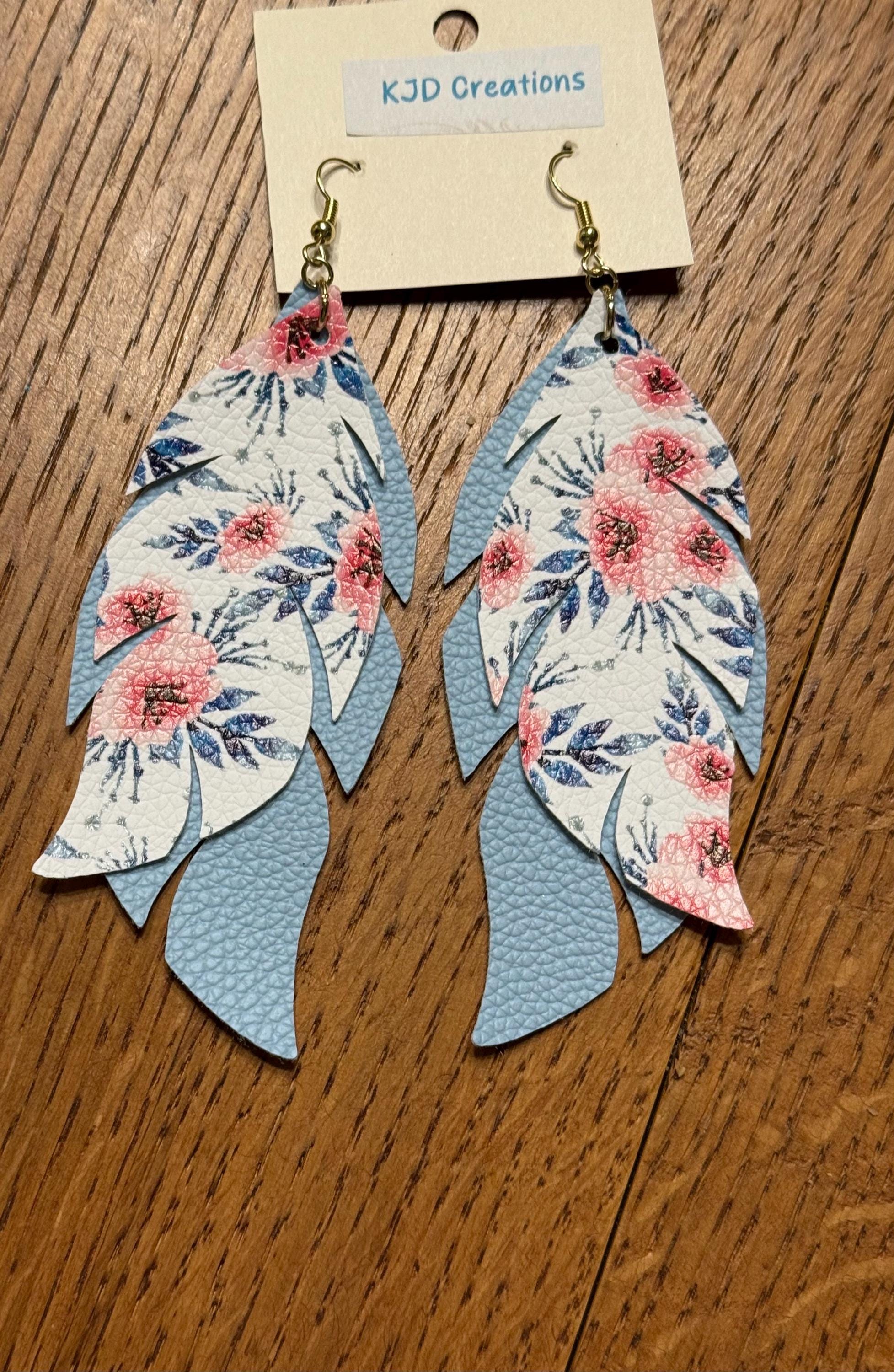 A pair of dangling feather-shaped earrings with a floral print on the front and light blue textured backing, hanging from gold hooks on a display card labeled KJD Creations, set against a wooden background.