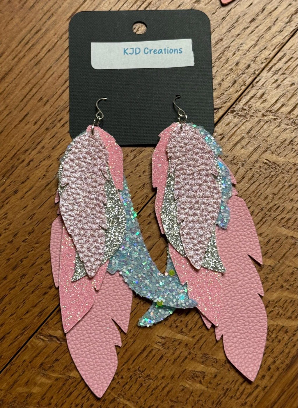 Pink feather-shaped dangle earrings with textured pink, glittery silver, and iridescent blue accents, displayed on a black card labeled KJD Creations over a wooden surface.