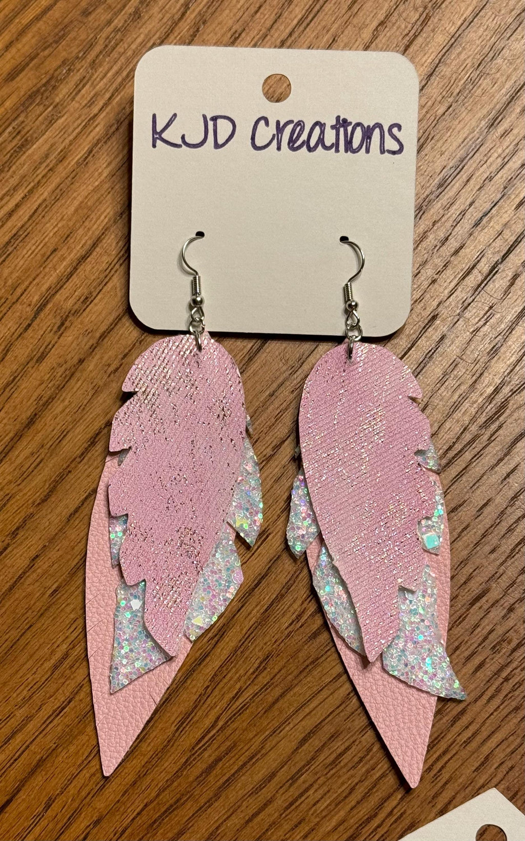 Pink feather-shaped dangle earrings with silver-toned hooks and iridescent glitter accents, displayed on a white card labeled KJD creations over a wooden surface.