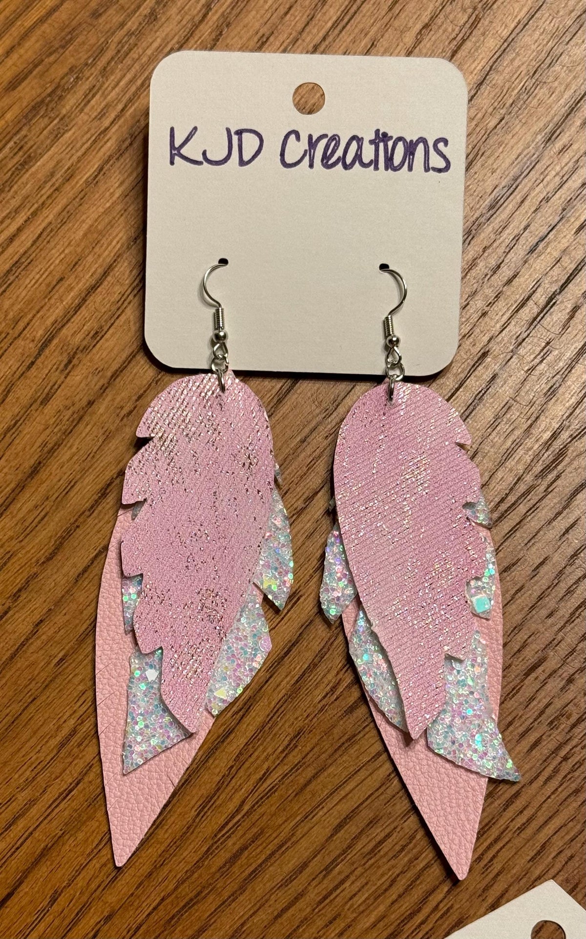 Pink feather-shaped dangle earrings with silver-toned hooks and iridescent glitter accents, displayed on a white card labeled KJD creations over a wooden surface.
