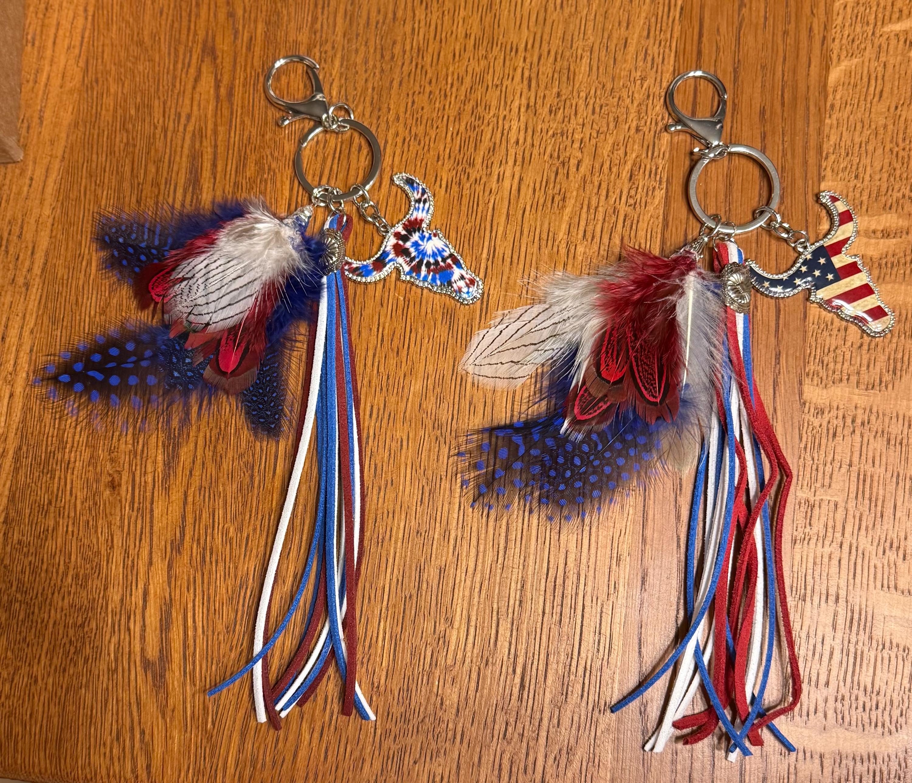Flat lay of both Western boho longhorn steer purse charm and keychain variants on wood — Tie Dye with red, white and blue tie-dye longhorn steer charm on left, Stars & Stripes with American flag longhorn steer charm on right — both with mixed feather bundles and red, white and blue suede fringe tassel strands
