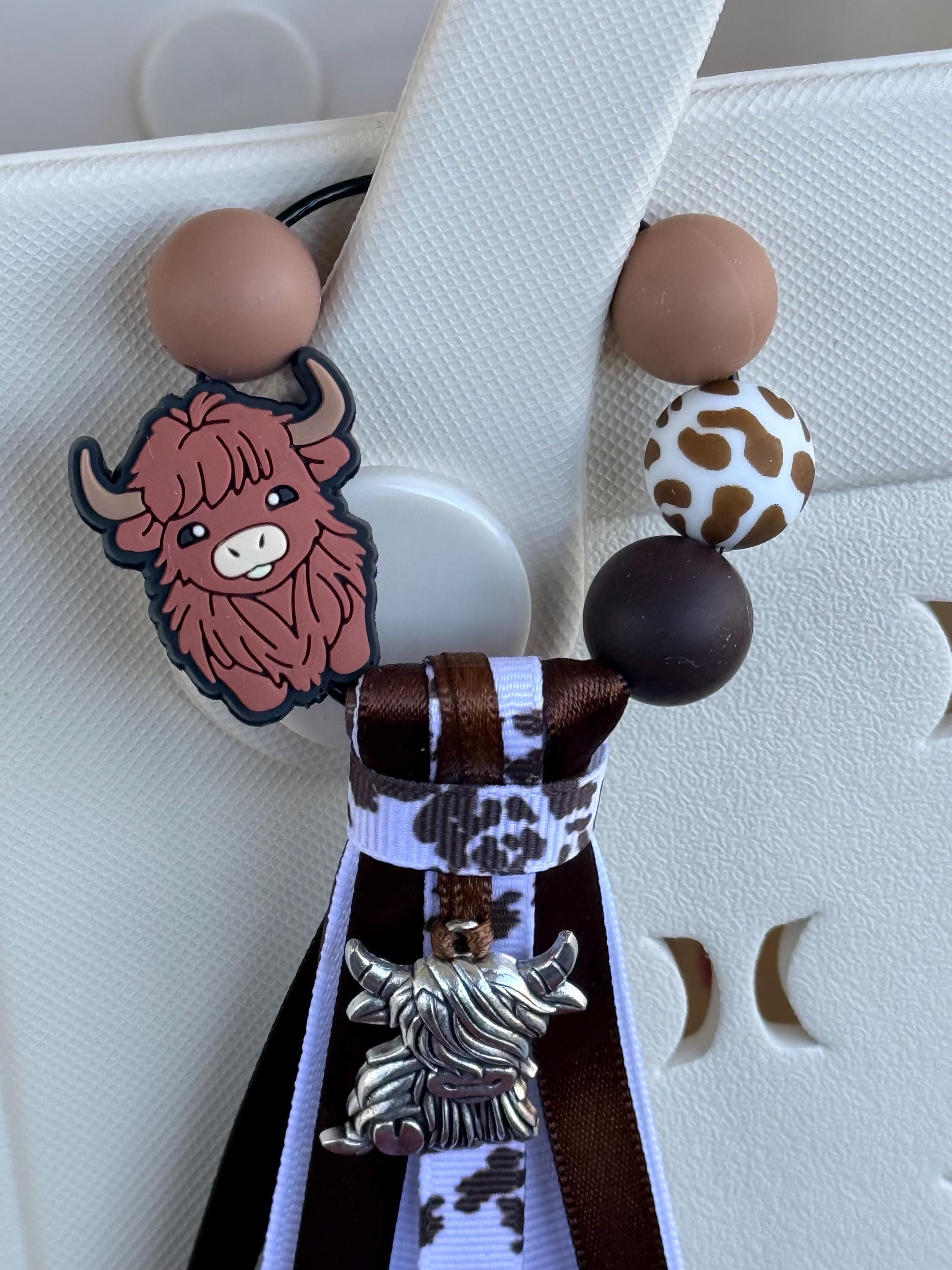 Highland Cow silicone beaded bag tassel with cow print ribbon, silver highland cow charm, and long brown and white ribbon tassel clipped onto a white tote bag handle
