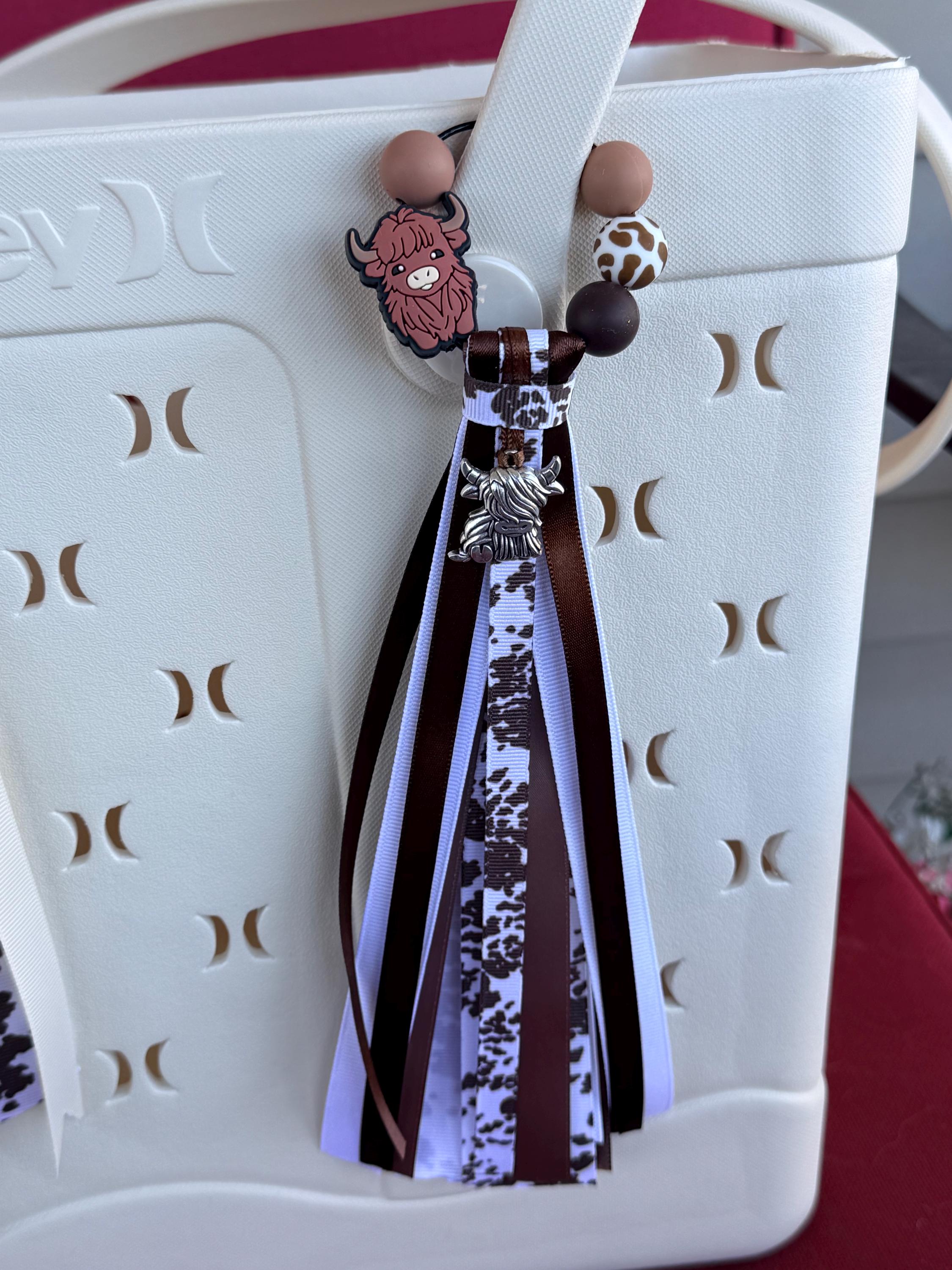 Highland Cow silicone beaded bag tassel with cow print ribbon, silver highland cow charm, and long brown and white ribbon tassel clipped onto a white tote bag handle
