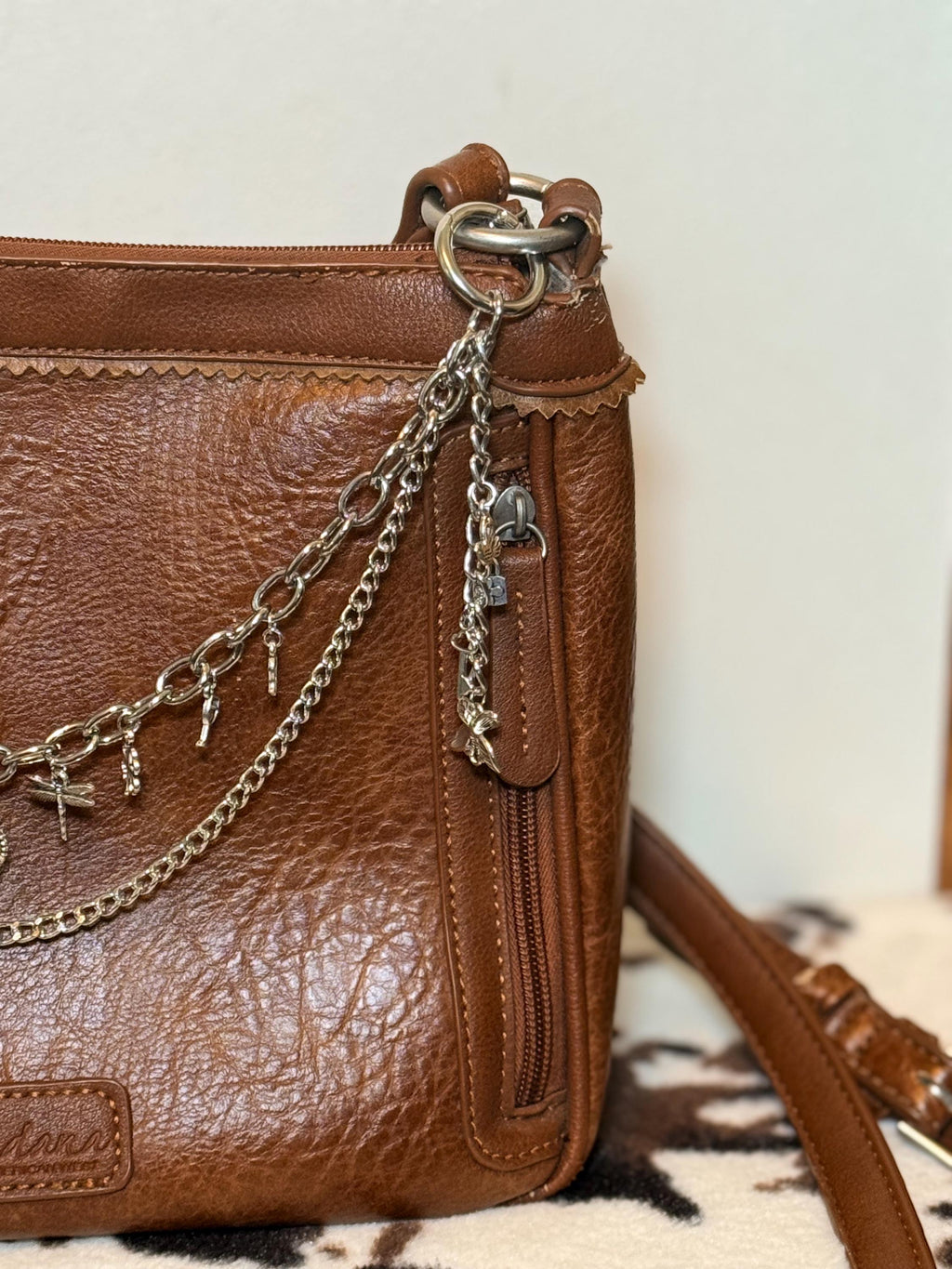 Close-up of right side hardware of tan distressed Bandana leather crossbody bag showing gold-tone oval link chain and delicate curb chain clipped to ring with key and padlock charms hanging from side hardware, Western boho charm purse chain detail