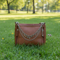 Front view of tan distressed Bandana leather crossbody bag on cowhide rug with two-strand gold-tone purse chain featuring large oval link chain and delicate curb chain with seven gold charms including four-leaf clover, butterfly, dragonfly, feather, arrow, key, and padlock, Western boho handbag jewelry