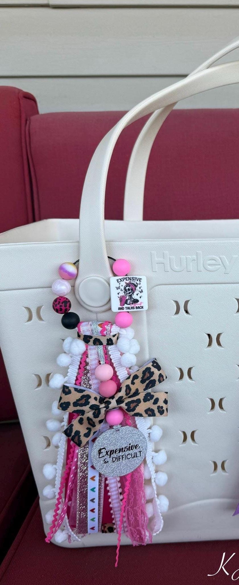 Close-up of Expensive Difficult & Talks Back silicone beaded bag tassel showing leopard bow, glitter charm disc, pink and black silicone beads, Expensive Difficult And Talks Back square bead, and layered ribbon tassel on a white Hurley bag
