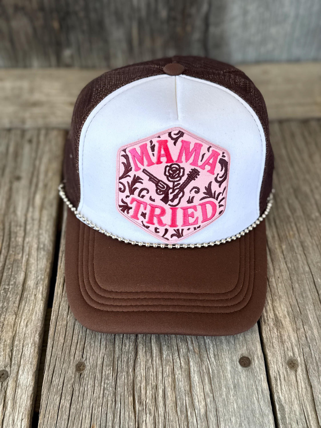 A brown and white trucker hat with a pink embroidered patch on the front that reads 'MAMA TRIED' with a rose and scroll design, displayed on a rustic wooden surface.