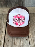 A brown and white trucker hat with a pink embroidered patch on the front that reads 'MAMA TRIED' with a rose and scroll design, displayed on a rustic wooden surface.