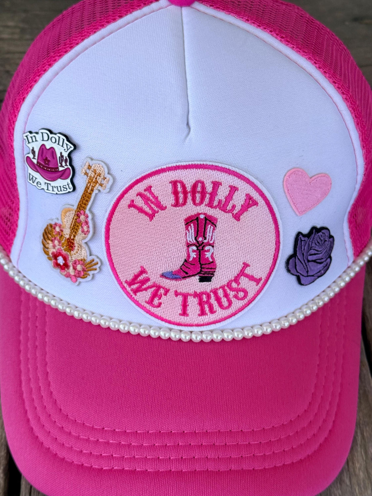 A close-up of a pink and white trucker hat featuring embroidered patches. The front panel is white with a pink brim and mesh back, decorated with a central circular patch with cowboy boots, a guitar patch, a pink heart, and a purple rose.
