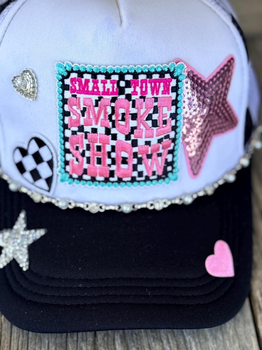 A close-up of a decorative trucker cap with a white crown, black brim, and multiple embellishments. The front pink letters over a black-and-white checkered background, bordered in turquoise rhinestones.