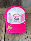 A bright pink and white trucker-style cap with embroidered patches, including a starfish embellishment on the brim, displayed on a rustic wooden surface.