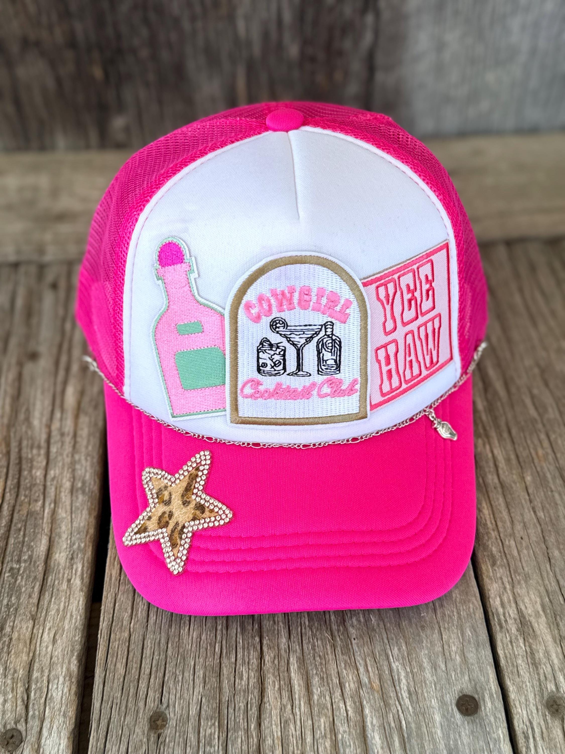 A bright pink and white trucker-style cap with embroidered patches, including a starfish embellishment on the brim, displayed on a rustic wooden surface.