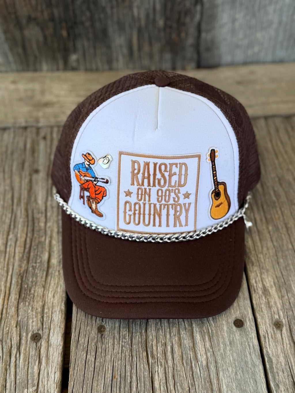 A brown and white trucker hat with a graphic that reads 'RAISED ON 90'S COUNTRY' and features illustrations of a cowboy playing a guitar and an acoustic guitar, resting on a rustic wooden surface.