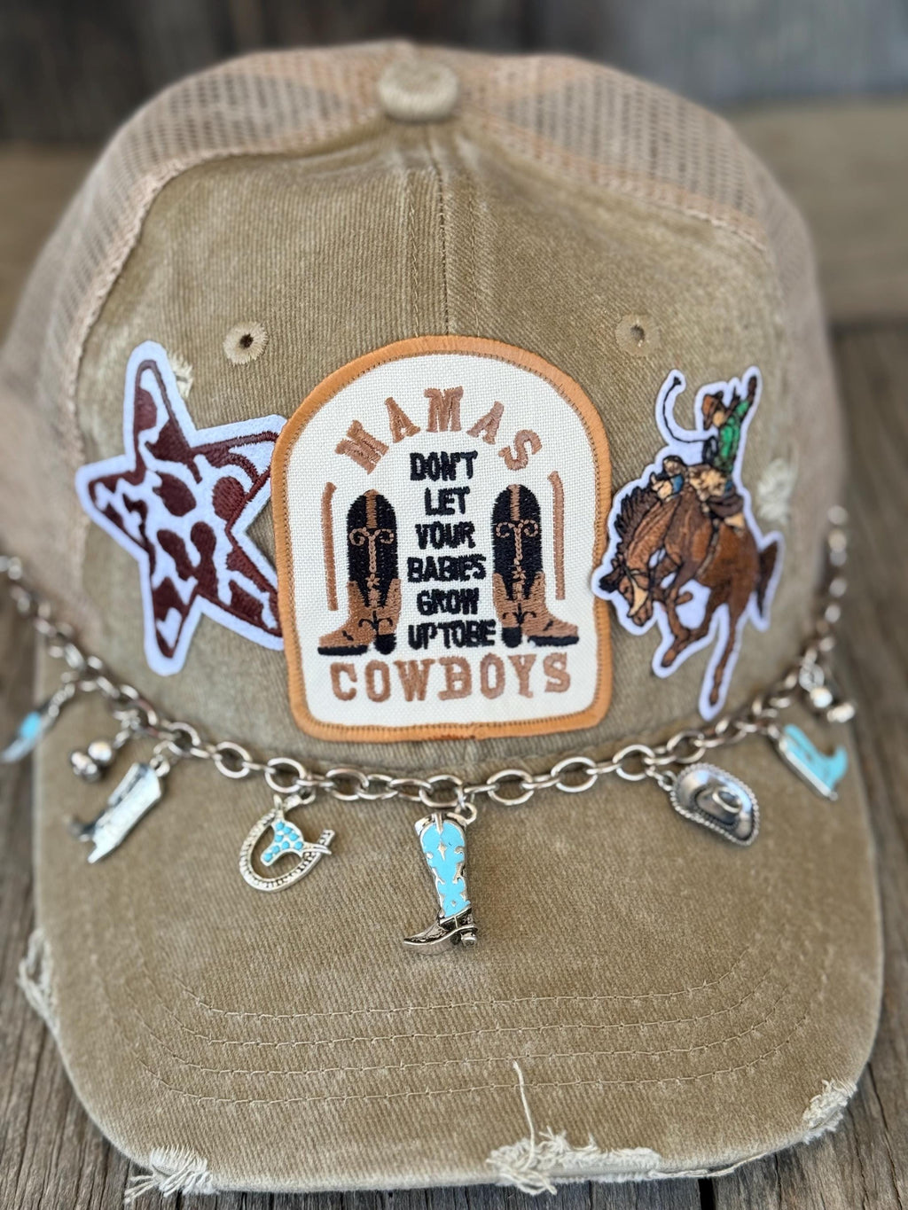 A distressed tan trucker cap with a western-themed embroidered patch cowboy and boot motifs, a star patch, and a silver charm bracelet draped across the brim.