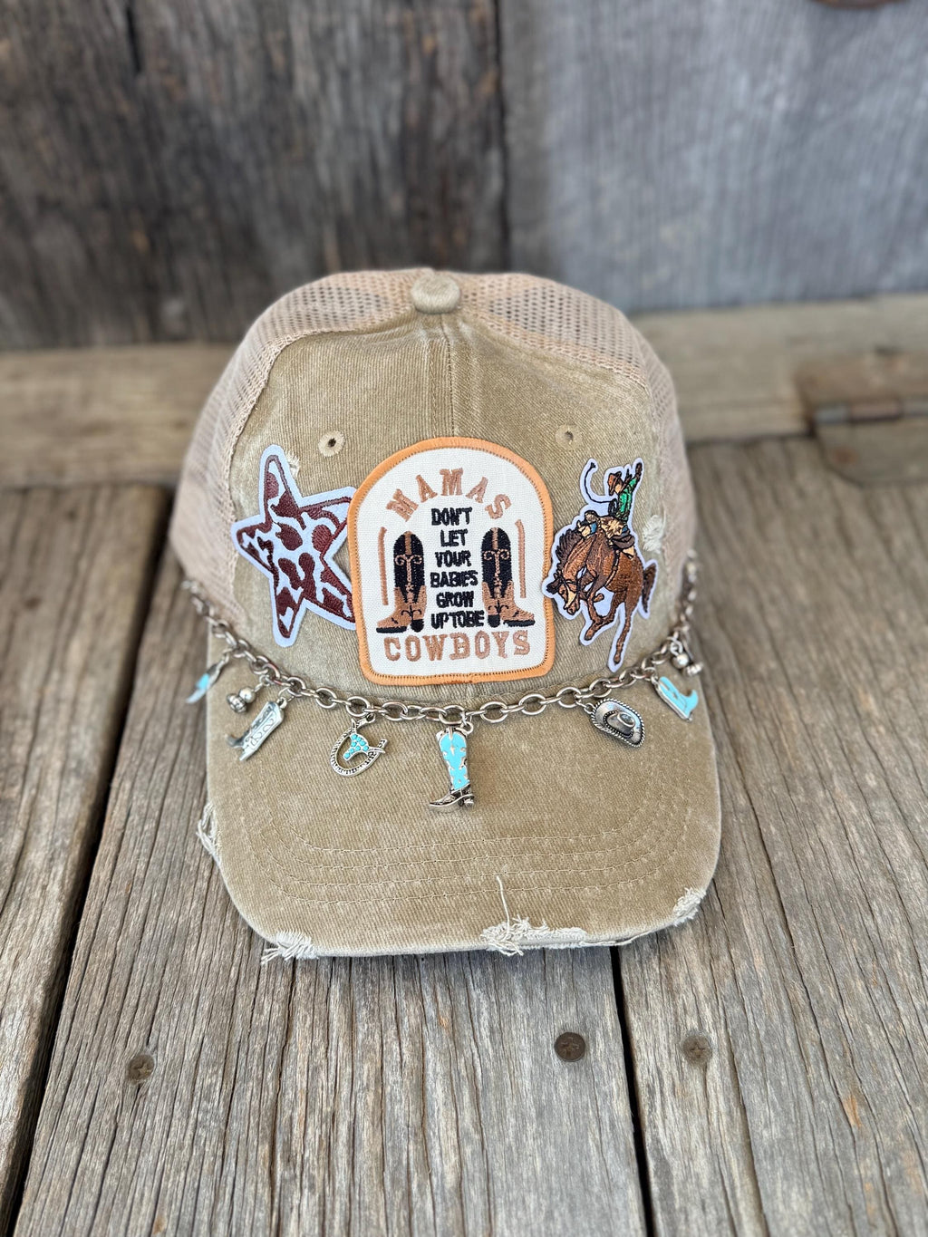A distressed tan trucker cap with a western 'Cowboys' theme, featuring embroidered patches, a decorative chain with cowboy boots charms, and a rustic wood plank background.