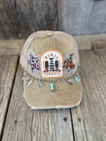 A distressed tan trucker cap with a western 'Cowboys' theme, featuring embroidered patches, a decorative chain with cowboy boots charms, and a rustic wood plank background.