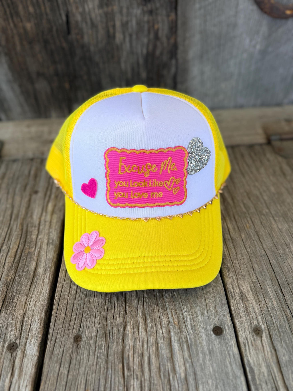 A yellow and white trucker hat with a pink embroidered patch and decorative pink and silver heart embellishments, displayed on a rustic wooden surface.